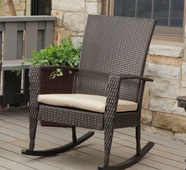 A brown wicker rocking chair with a beige cushion sits on a wooden deck outside a stone house.